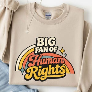 Op de afbeelding: Beige crewneck sweatshirt met de tekst "BIG FAN OF Human Rights" in een retro regenboogontwerp. De sweatshirt is opgevouwen met een spijkerbroek en een zilveren ketting.