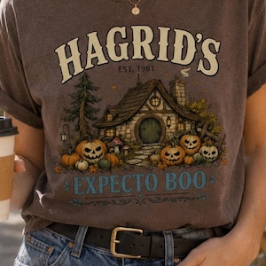 May include: A brown short-sleeve t-shirt featuring a graphic print of a cottage surrounded by pumpkins and trees. The text reads HAGRID'S EST. 1981 and EXPECTO BOO in a decorative font. The shirt has a crew neckline and rolled sleeves.