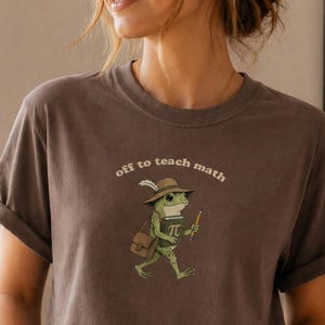 May include: A brown t-shirt featuring the phrase "off to teach math" and a cartoon frog illustration. The frog is depicted wearing a hat, carrying a bag, and holding a pencil, with a pi symbol on its chest. The shirt is worn with blue jeans.