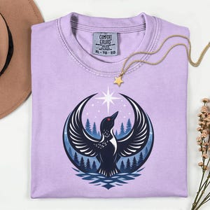 May include: A light purple t-shirt featuring a graphic of a black bird with spread wings, a red eye, and a white star above it, set against a backdrop of blue pine trees. The shirt has a Comfort Colors label with size XL, TG, EG printed inside.