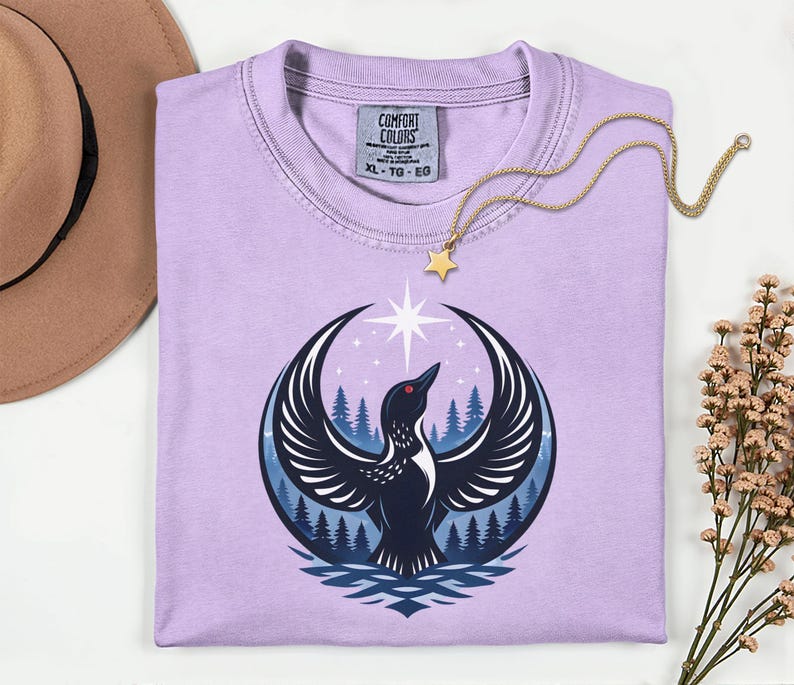 May include: A light purple t-shirt featuring a graphic of a black bird with spread wings, a red eye, and a white star above it, set against a backdrop of blue pine trees. The shirt has a Comfort Colors label with size XL, TG, EG printed inside.
