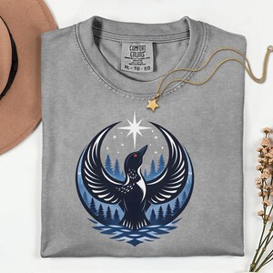 May include: A grey t-shirt with a Comfort Colors label in the neck, featuring a graphic of a black loon with wings spread wide, a bright white star above, and a blue forest background. A gold chain with a small star pendant rests on the shirt.