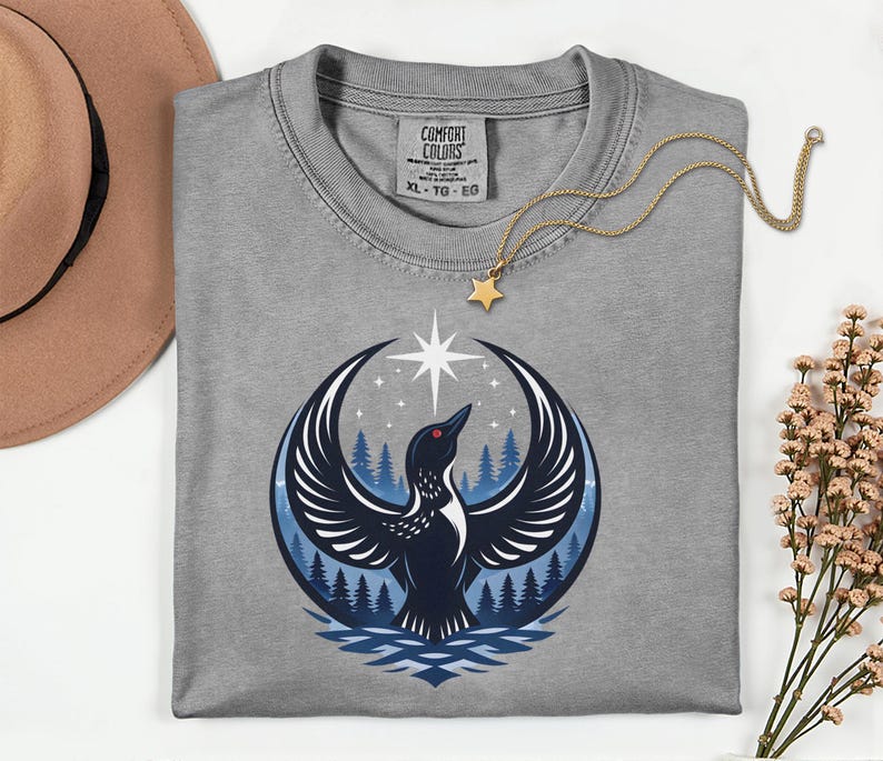 May include: A grey t-shirt with a Comfort Colors label in the neck, featuring a graphic of a black loon with wings spread wide, a bright white star above, and a blue forest background. A gold chain with a small star pendant rests on the shirt.