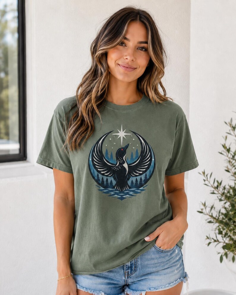 May include: An olive green short-sleeve t-shirt featuring a graphic print of a black bird with spread wings, positioned in front of a dark blue forest and a crescent moon shape, topped with a white four-pointed star against a light background.
