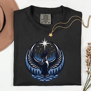 May include: A black crewneck t-shirt featuring a graphic of a loon with wings spread wide against a dark blue forest and night sky background with a bright star. The shirt has a Comfort Colors label in size XL, TG, EG and a gold star pendant necklace.