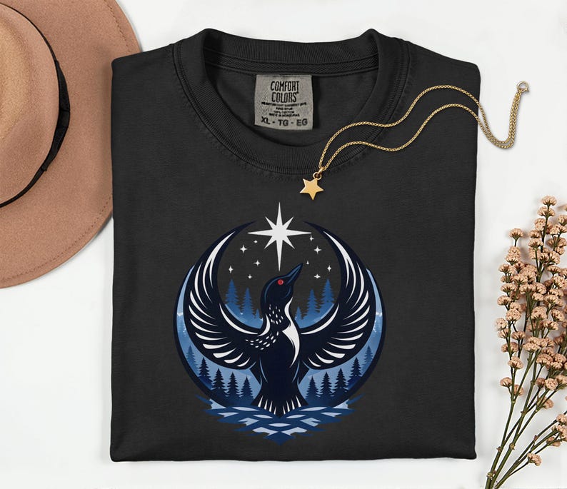 May include: A black crewneck t-shirt featuring a graphic of a loon with wings spread wide against a dark blue forest and night sky background with a bright star. The shirt has a Comfort Colors label in size XL, TG, EG and a gold star pendant necklace.