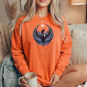 May include: An orange long-sleeve crewneck sweatshirt featuring a graphic print of a black loon with wings spread wide, positioned above a blue water ripple design and a dark forest silhouette, topped with a white four-pointed star.