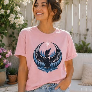 May include: A light pink short-sleeve t-shirt featuring a graphic print of a black loon with wings spread wide, positioned in front of a dark blue forest and water scene, topped with a white four-pointed star against a circular background.