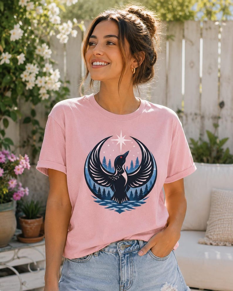 May include: A light pink short-sleeve t-shirt featuring a graphic print of a black loon with wings spread wide, positioned in front of a dark blue forest and water scene, topped with a white four-pointed star against a circular background.