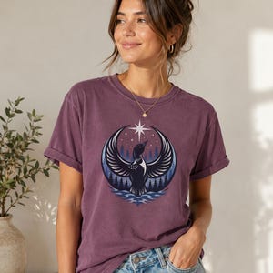May include: A plum-colored short-sleeve t-shirt featuring a circular graphic print of a loon with wings spread wide, positioned above stylized water and a forest of pine trees under a night sky with a prominent white star at the top center.