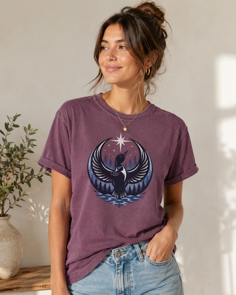 May include: A plum-colored short-sleeve t-shirt featuring a circular graphic print of a loon with wings spread wide, positioned above stylized water and a forest of pine trees under a night sky with a prominent white star at the top center.
