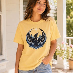 May include: A pale yellow short-sleeve t-shirt featuring a graphic print of a black loon with wings spread wide, positioned in front of a dark blue forest and a bright white star, all contained within a circular frame above stylized blue water.