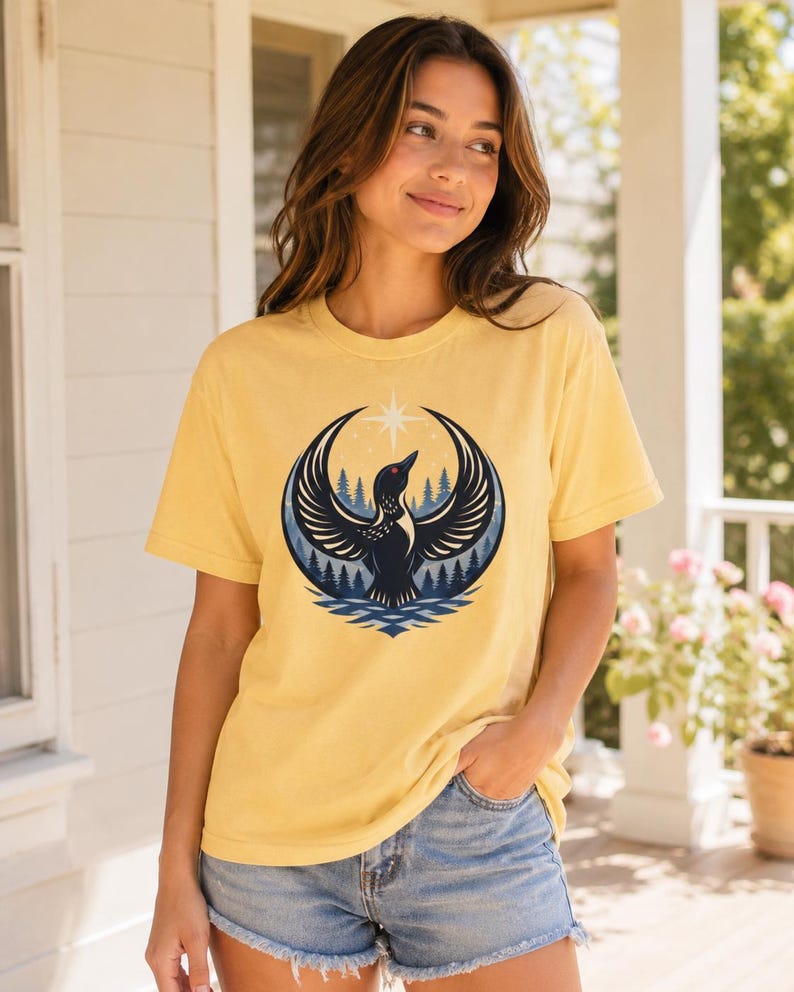 May include: A pale yellow short-sleeve t-shirt featuring a graphic print of a black loon with wings spread wide, positioned in front of a dark blue forest and a bright white star, all contained within a circular frame above stylized blue water.