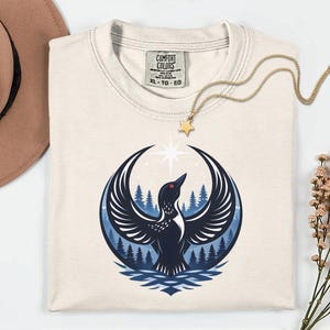 May include: A cream-colored t-shirt with a Comfort Colors label in size XL. The graphic features a black bird with wings spread wide, positioned in front of a blue forest and a bright white star, all contained within a circular design.