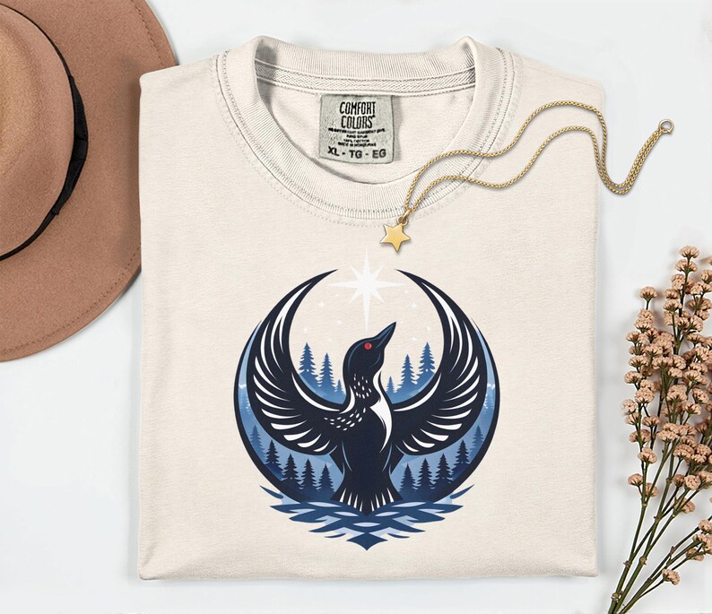 May include: A cream-colored t-shirt with a Comfort Colors label in size XL. The graphic features a black bird with wings spread wide, positioned in front of a blue forest and a bright white star, all contained within a circular design.