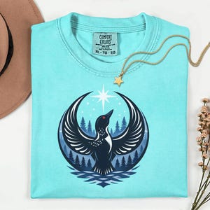 May include: A light blue short-sleeve t-shirt featuring a graphic of a black bird with wings spread wide, a forest background, and a bright star above. The inner neck label displays the text Comfort Colors with size XL, TG, EG.