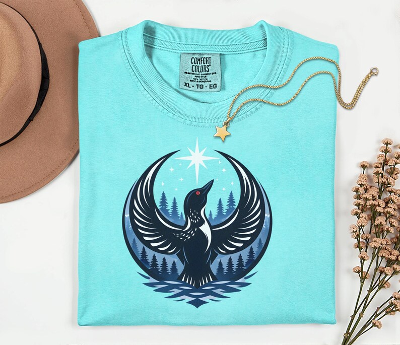May include: A light blue short-sleeve t-shirt featuring a graphic of a black bird with wings spread wide, a forest background, and a bright star above. The inner neck label displays the text Comfort Colors with size XL, TG, EG.