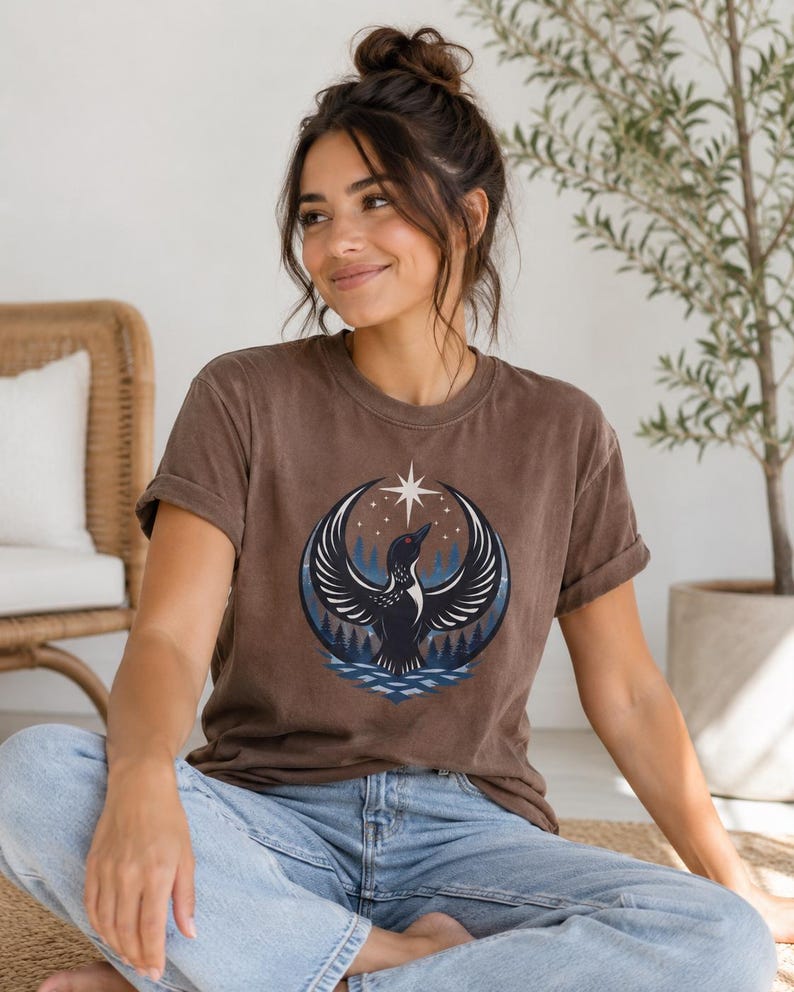 May include: A brown short-sleeve t-shirt featuring a graphic print of a black bird with wings spread wide, positioned above a blue water pattern and a dark forest silhouette, all framed by a crescent shape and a single white star above.