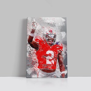 May include: A canvas print of a football player in a red uniform with the number 2. The player is wearing a helmet and raising his arm. The background has a gray and white splatter effect.