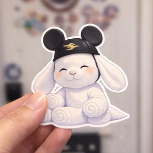 Puede incluir: Una pegatina de un conejito blanco con un sombrero negro con un emblema dorado. El conejito tiene una cara sonriente y mejillas rosadas. La pegatina está sujeta por una mano, con un fondo borroso.