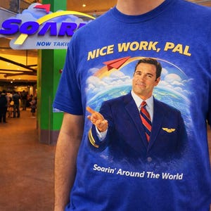 May include: Blue t-shirt with the text "NICE WORK, PAL" and an image of a man in a pilot's uniform. The image also includes a paper airplane and the words "Soarin' Around The World".