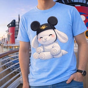 May include: Pale blue t-shirt with a cartoon bunny wearing a black hat with Mickey Mouse ears. The white bunny has pink cheeks and is sitting down. The shirt is being worn on a cruise ship deck.