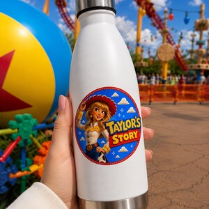 May include: A white stainless steel water bottle with a silver cap and base. A circular sticker on the front features a cartoon cowgirl character wearing a red hat and the text Taylor's Story against a blue background with white clouds.