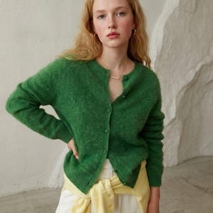 May include: A green cardigan with a round neckline and button closure. The model is wearing a gold necklace and hoop earrings. The cardigan is paired with white pants and a yellow sweater tied around the waist. The background is a neutral color.