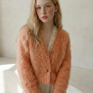 May include: A peach-colored, fuzzy cardigan with a button-up front. The cardigan is paired with a cream-colored lace-trimmed top and a silver necklace. The model has long blonde hair and is wearing silver earrings.