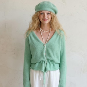 Puede incluir: Un conjunto de boina y cárdigan verde menta. El cárdigan tiene cuello en V y cintura con cordón. Se lleva un collar largo de perlas sobre el cárdigan. El conjunto se completa con pantalones blancos.