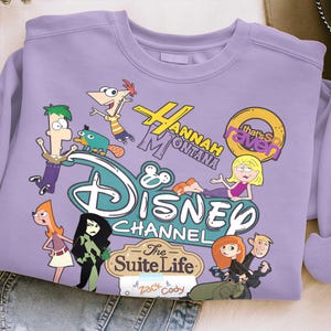 May include: A lilac sweatshirt with a Disney Channel graphic. The design features characters and logos from "Hannah Montana," "That's So Raven," and "The Suite Life of Zack & Cody." The graphic includes cartoon characters and the Disney Channel logo.