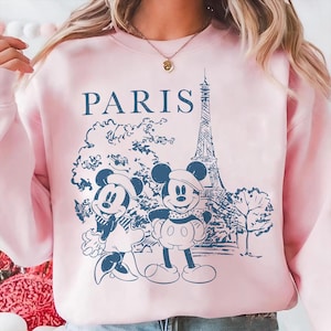 K&ouml;nnte beinhalten: Hellrosa Sweatshirt mit einer blauen Illustration von Mickey und Minnie Mouse in Paris, mit dem Eiffelturm und dem Wort "PARIS". Das Design hat einen Vintage-Skizzenstil.