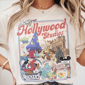 Puede incluir: Camiseta color crema con un gráfico colorido de Disney's Hollywood Studios. El diseño incluye varios personajes, las palabras "Hollywood Studios" y "Rockers". La camiseta tiene cuello redondo y mangas cortas.