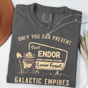 May include: Dark gray Comfort Colors t-shirt with the text "ONLY YOU CAN PREVENT" and "Visit ENDOR Lunar Forest". The shirt features a cartoon Ewok character holding a shovel. A silver necklace with a pendant is draped across the shirt.