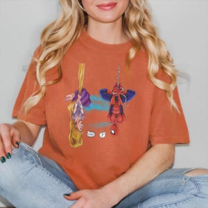May include: Burnt orange t-shirt featuring a cartoon illustration of Rapunzel and Spider-Man hanging upside down. Rapunzel is holding onto her long blonde hair, and Spider-Man is hanging from a web. The shirt has the text "Oh Hai!" and a question mark.