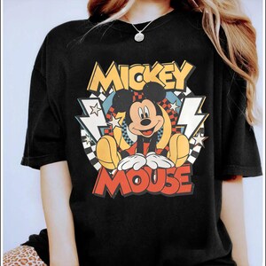 K&ouml;nnte beinhalten: Schwarzes T-Shirt mit einem farbenfrohen Mickey Mouse-Motiv. Das Design enth&auml;lt die Worte "Mickey Mouse" in einem Retro-Schriftstil, zusammen mit Sternen, Blitzen und einem Schachbrettmuster. Das T-Shirt hat einen klassischen Rundhalsausschnitt.