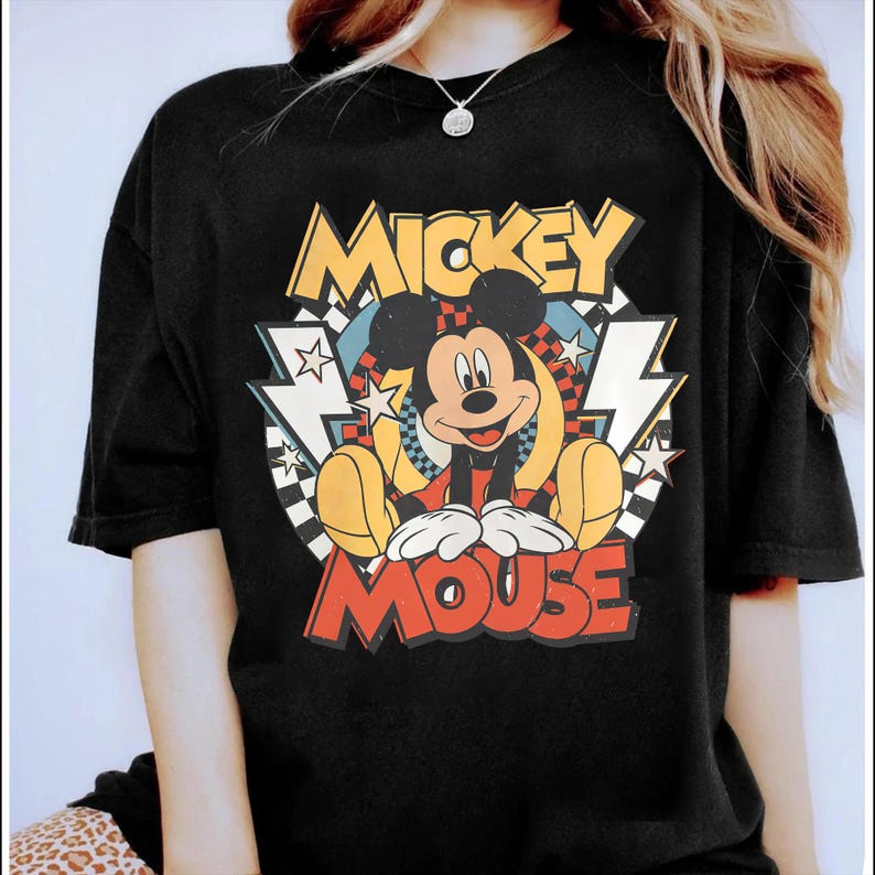 K&ouml;nnte beinhalten: Schwarzes T-Shirt mit einem farbenfrohen Mickey Mouse-Motiv. Das Design enth&auml;lt die Worte "Mickey Mouse" in einem Retro-Schriftstil, zusammen mit Sternen, Blitzen und einem Schachbrettmuster. Das T-Shirt hat einen klassischen Rundhalsausschnitt.