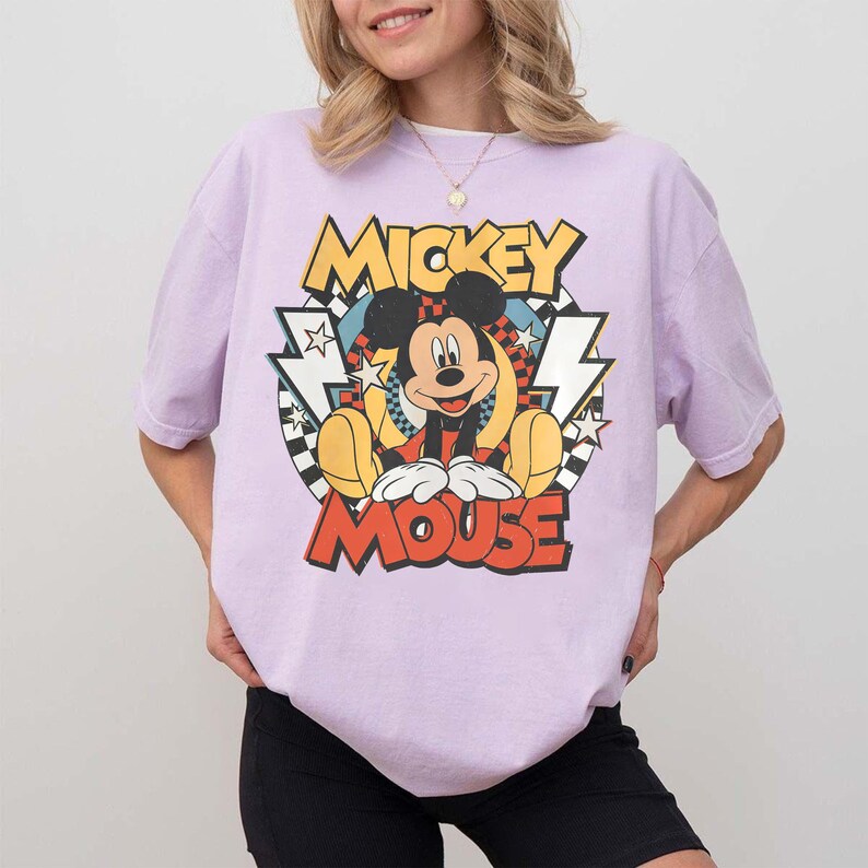 K&ouml;nnte beinhalten: Helllila T-Shirt mit gro&szlig;em Mickey Mouse-Motiv. Das Design enth&auml;lt die Worte "Mickey Mouse" in einem Retro-Stil, mit Mickey Mouse in der Mitte. Das T-Shirt hat kurze &Auml;rmel.