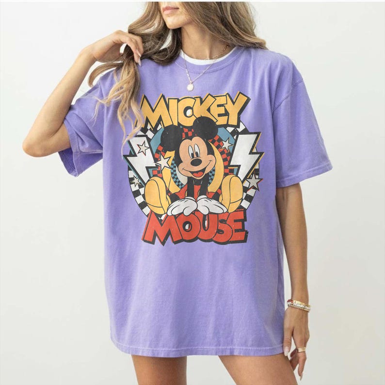 K&ouml;nnte beinhalten: Ein lila T-Shirt mit einem Mickey Mouse-Motiv. Das Design zeigt die Worte "Mickey Mouse" und Mickey in sitzender Pose. Das T-Shirt hat kurze &Auml;rmel und eine lockere Passform. Das T-Shirt ist helllila.