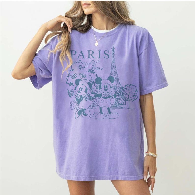 K&ouml;nnte beinhalten: Ein lila T-Shirt mit einer Grafik von Mickey und Minnie Mouse vor dem Eiffelturm, mit dem Wort "PARIS" dar&uuml;ber. Das T-Shirt hat kurze &Auml;rmel und einen Rundhalsausschnitt.