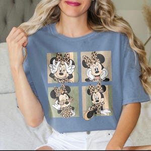 Puede incluir: Una camiseta azul con un diseño de cuatro paneles de Minnie Mouse en varias poses. Cada Minnie Mouse tiene un lazo y un atuendo con estampado de leopardo. El fondo de cada panel tiene un diseño de rayas beige y verde.