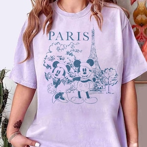 K&ouml;nnte beinhalten: Helllila T-Shirt mit blauem Aufdruck von Mickey und Minnie Mouse in Paris, mit dem Eiffelturm im Hintergrund. Das Wort "PARIS" ist &uuml;ber dem Motiv aufgedruckt.