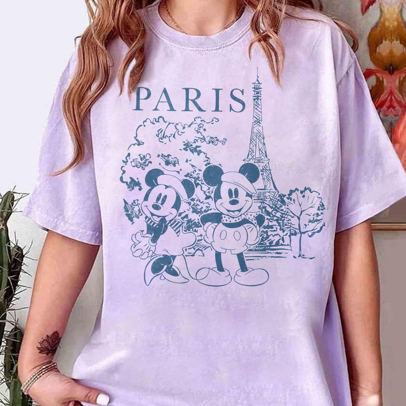 K&ouml;nnte beinhalten: Helllila T-Shirt mit blauem Aufdruck von Mickey und Minnie Mouse in Paris, mit dem Eiffelturm im Hintergrund. Das Wort "PARIS" ist &uuml;ber dem Motiv aufgedruckt.
