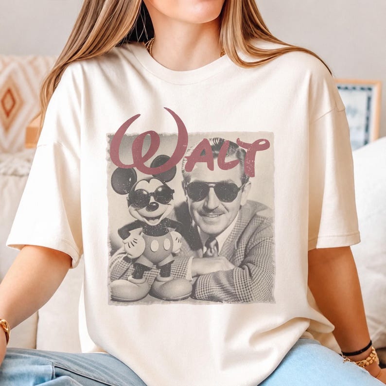 K&ouml;nnte beinhalten: Ein cremefarbenes Kurzarm-T-Shirt mit einem Grafikdruck von Walt Disney und Mickey Mouse, die beide eine Sonnenbrille tragen. Der Text "WALT" ist in einer gro&szlig;en, stilisierten kastanienbraunen Schrift &uuml;ber dem schwarz-wei&szlig;en Fotobild gedruckt.