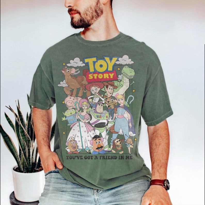 May include: Green t-shirt featuring a colorful graphic of characters from the animated film "Toy Story." The design includes the words "Toy Story" in yellow and red, and the phrase "You've Got a Friend in Me." The shirt is worn by a person.