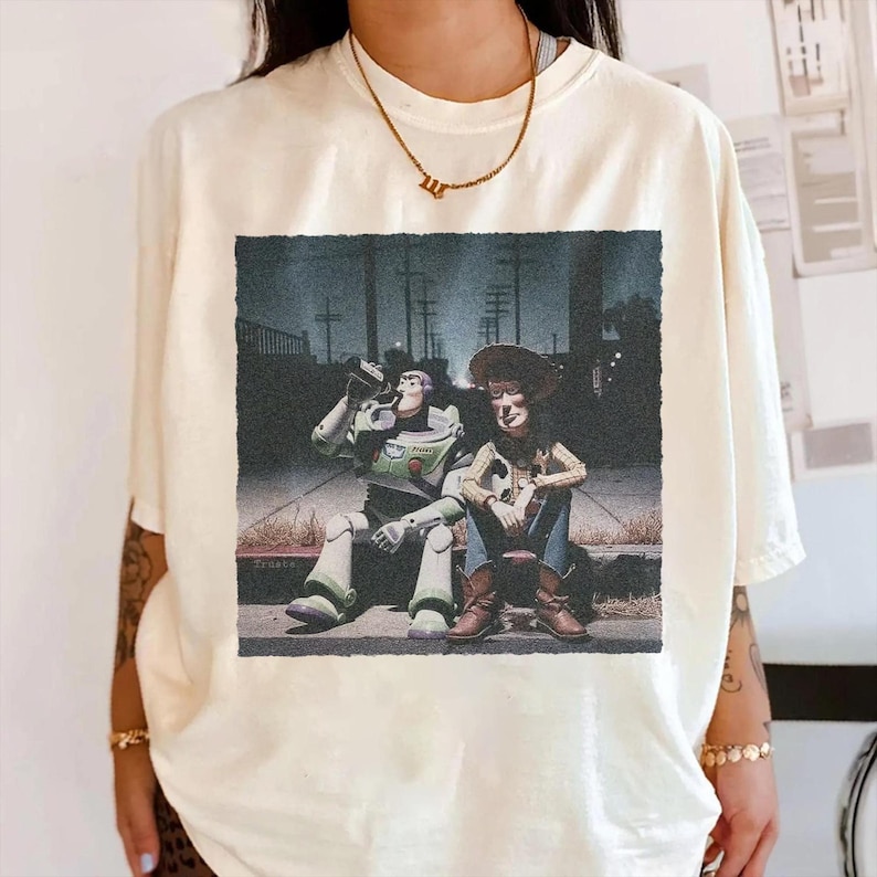 May include: Cream-colored t-shirt featuring a graphic of Buzz Lightyear and Woody from Toy Story. The image shows the characters sitting on a curb in a nighttime setting. The shirt has short sleeves and a relaxed fit.