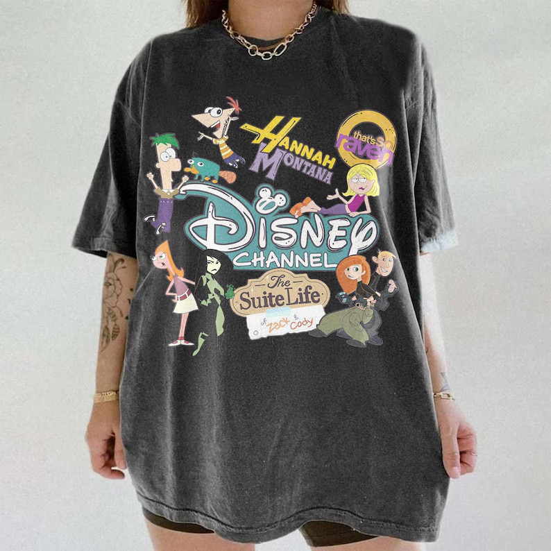 May include: Dark grey t-shirt with a Disney Channel graphic featuring characters from shows such as Hannah Montana, Phineas and Ferb, and The Suite Life of Zack & Cody. The graphic is in various colours, including blue, green, and yellow.