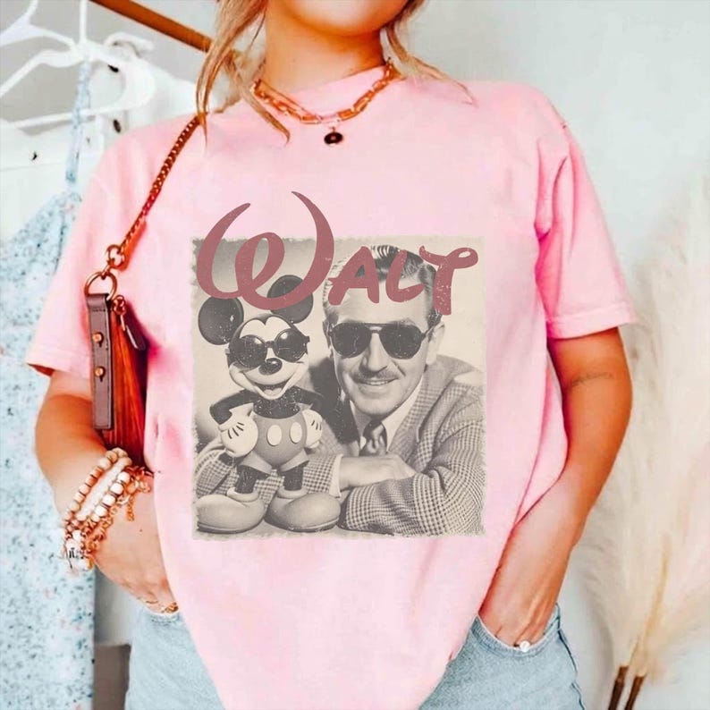 K&ouml;nnte beinhalten: Ein hellrosa T-Shirt mit kurzen &Auml;rmeln und einem Grafikdruck von Walt Disney und Mickey Mouse, die beide eine Sonnenbrille tragen. Der Text "WALT" ist in einer gro&szlig;en, stilisierten Schriftart &uuml;ber den Figuren aufgedruckt.