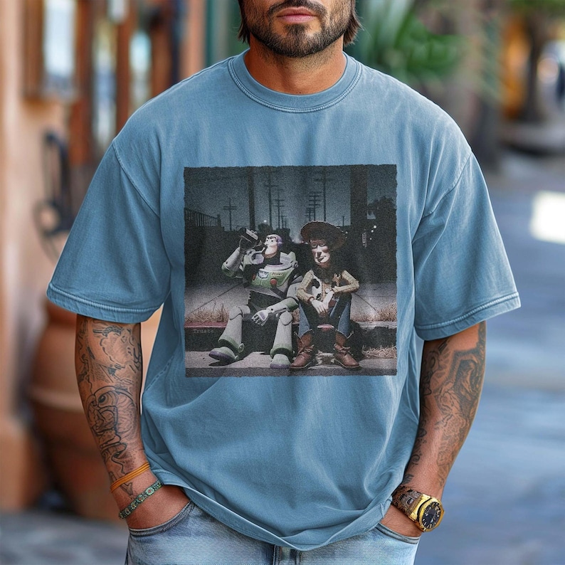 May include: A light blue t-shirt featuring a graphic of two animated characters sitting on a curb. One character is a space ranger, the other is a cowboy. The graphic is in a rectangular frame.