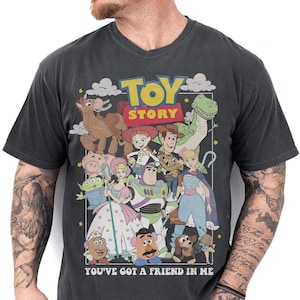 May include: Dark gray v-neck t-shirt featuring a colorful graphic of characters from the animated film "Toy Story." The design includes Woody, Buzz Lightyear, and other beloved characters, with the text "TOY STORY" and "YOU'VE GOT A FRIEND IN ME."